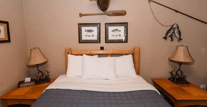 a Bed with wooden headboard, white pillows, grey blanket, two lamps, framed art, and fishing themed decor on the wall in a room at Shaver Lake Village Hotel.