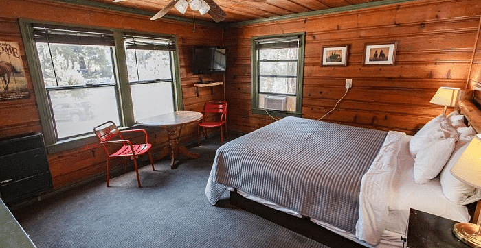 a Cozy bedroom at Shaver Lake Village Hotel with wood paneled walls, two windows, a double bed, and red chairs next to a small table.