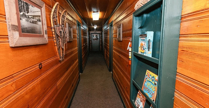 a Hallway at Shaver Lake Village Hotel with wood paneling, artwork on the walls, and a green bookshelf on the right side, leading to rooms.