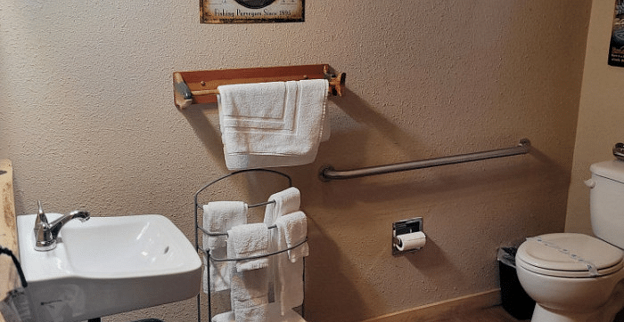 a Bathroom at Shaver Lake Village Hotel with a white sink, toilet, stacked white towels, grab bar, and a framed picture on the wall.