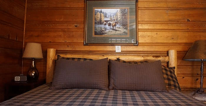 Close-up of a bed at Shaver Lake Village Hotel with a plaid comforter, brown pillows, and framed artwork above the wooden headboard.