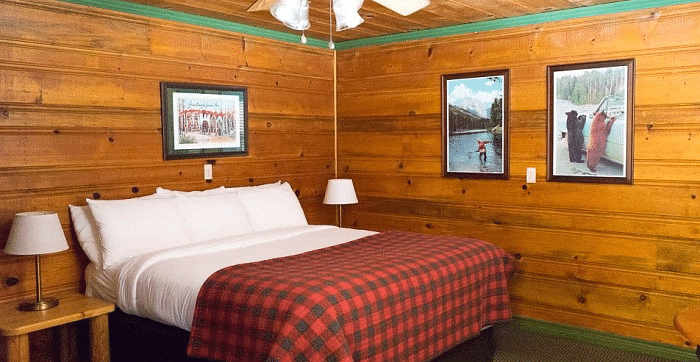 a Bedroom at Shaver Lake Village Hotel with wooden walls, plaid comforter, white pillows, framed artwork, and a ceiling fan.