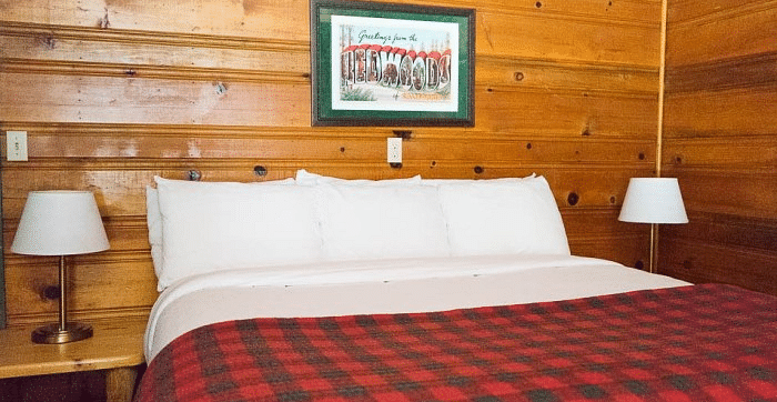 a Bed at Shaver Lake Village Hotel with plaid comforter, white pillows, wooden walls, framed art, and two bedside lamps.