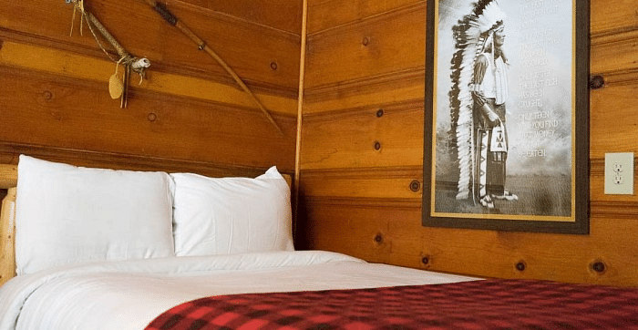 Overview of a Bed in a room at Shaver Lake Village Hotel with white pillows, red plaid blanket, wood paneled walls, framed art, and decorative sticks.