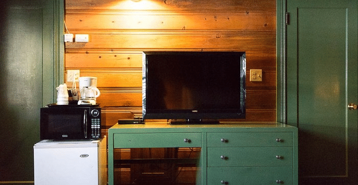 Overview of a Room at Shaver Lake Village Hotel with green dresser, TV, mini fridge, microwave, wood paneling, and two green doors.