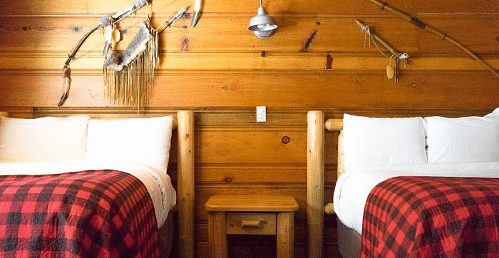 Overview of a room with Twin beds at Shaver Lake Village Hotel with red plaid blankets, white pillows, wooden headboards, and fishing gear decor on wood walls.