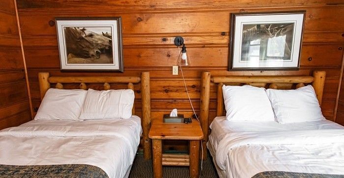 Twin beds in a room at Shaver Lake Village Hotel with white linens, wooden headboards, a small table, and framed artwork on wood walls.