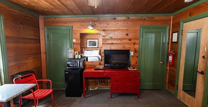 furniture inside a Room at Shaver Lake Village Hotel including a red desk, mini fridge, TV, two green doors, wood paneling, and red chairs.