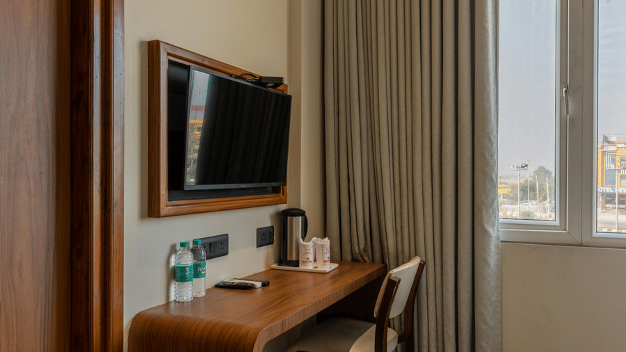 Wall-mounted TV placed in a corner of the room with a work desk right below, and a large glass window beside it with the curtains drawn in a Deluxe Room at Hotel Highway Prince, Behror.
