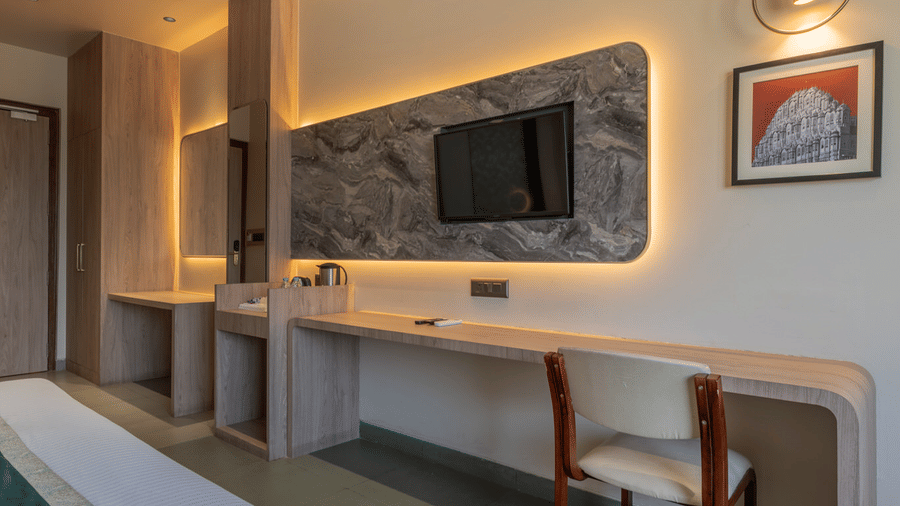 Spacious layout of Super Deluxe Room showcasing the plush double bed with pillows and cushions facing the wall-mounted TV placed on a panel, an a frosted glass bathroom door.