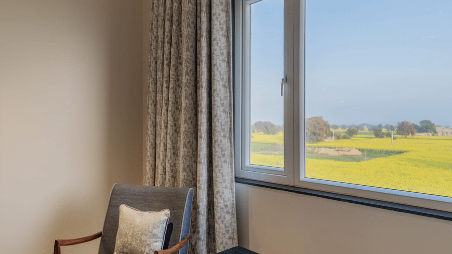 Hotel Highway Prince, Behror guest room corner showing a dark wood coffee table, a grey armchair, and a window with a view of green fields.