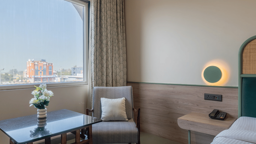 Seating area with an upholstered chairs and coffee table with a flower vase placed beside the double bed in a Super Deluxe Room at Hotel Highway Prince, Behror.