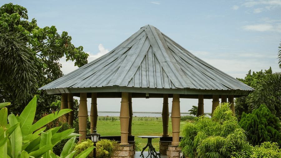 A wooden octagonal pavilion at Rhino & River Wildlife Retreat & Spa, Pobitora, framed by bright green foliage with a view of the open landscape in the background.