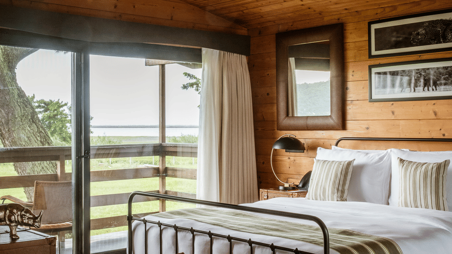 A vertical interior view of a room at Rhino & River Wildlife Retreat & Spa, Pobitora, showcasing the dark tiled floor, area rug, and metal bed frame near the balcony entrance.