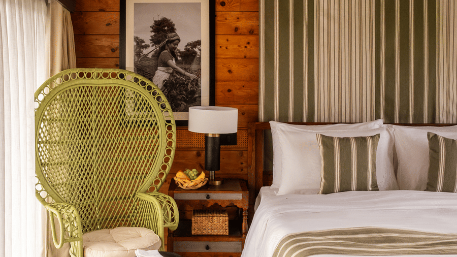 A close-up  view of a bedroom at Rhino & River Wildlife Retreat & Spa, Pobitora, showcasing a white bed, striped canopy, wicker chair, and large windows with white curtains.