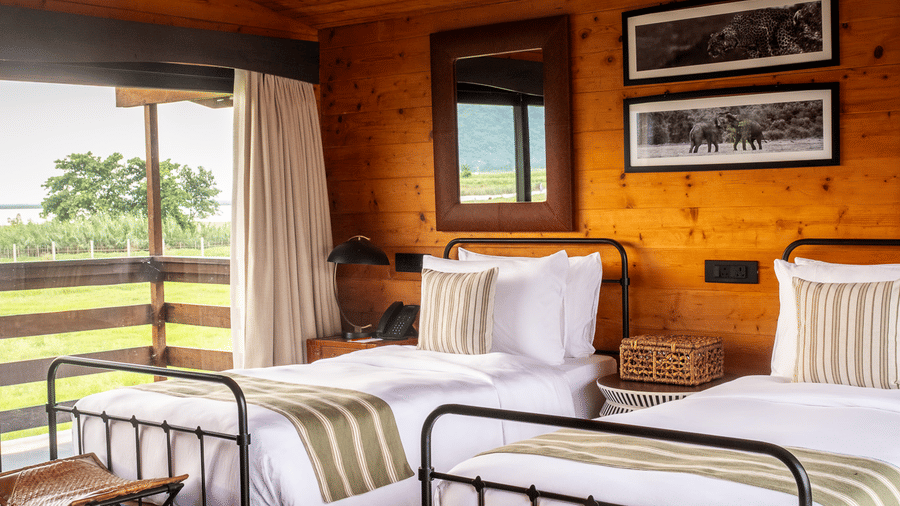 A vertical view of a twin bedroom at Rhino & River Wildlife Retreat & Spa, Pobitora, showcasing polished wooden ceilings, walls, and small leather folding stools at the foot of each bed.