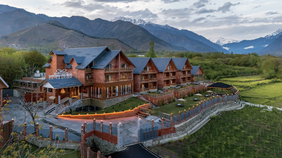Facade of Mountnoor Sarovar Premiere resort in Pahalgam, showing the long, multi-story timber and stone architecture nestled against a backdrop of valleys and towering, misty mountains.