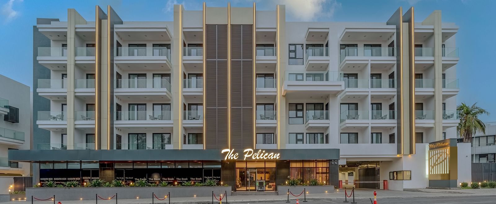 A tall white hotel building with palm trees in front and a clear blue sky in the background - The Pelican Hotel, Accra - Accra Hotel Images.