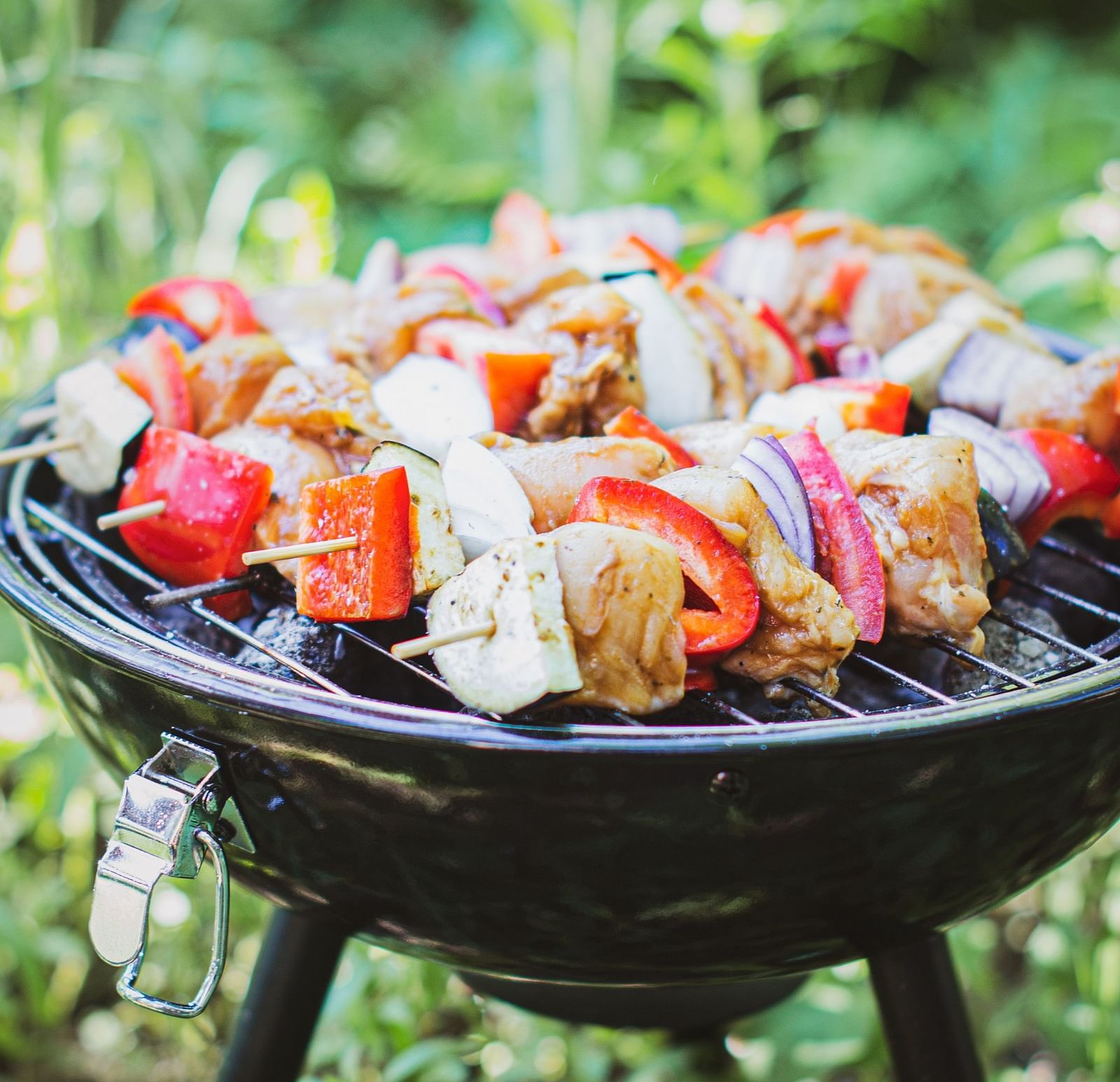 A barbecue grill set up in an open space, featuring skewers of vegetables and meat cooking over the flames
