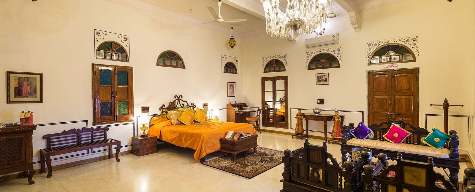 A well-lit bedroom at Fort Barli, featuring a vibrant orange bedspread, a crystal chandelier, and antique furniture, offers a spacious and elegant retreat.