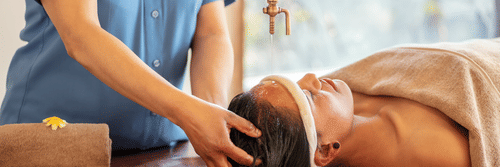 Woman receiving a traditional Ayurvedic head massage with warm oil being poured onto her forehead, administered by a therapist in a spa setting.  