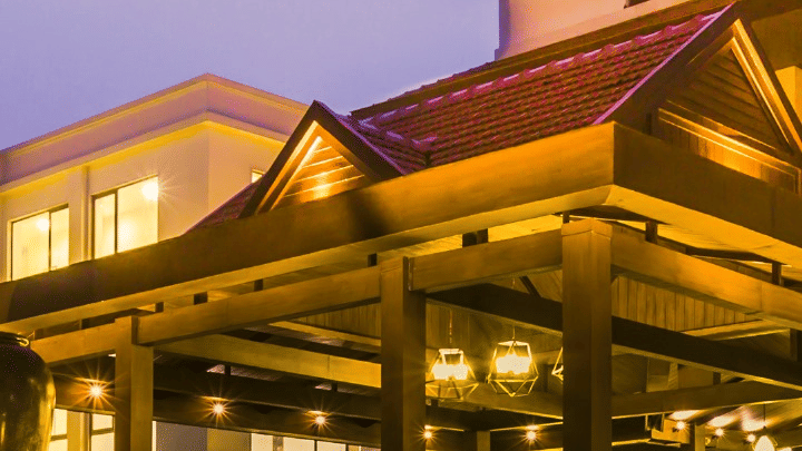 Exterior view of a resort or hotel building at dusk, featuring a large wooden portico entrance with a tiled roof, and tree branches against a twilight sky.