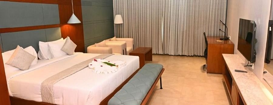 A hotel room at Dhanalakshmi Srinivasan Hotels, Perambalur, featuring a double bed, two benches, a TV on a console, a desk with a chair, two armchairs, and closed curtains.