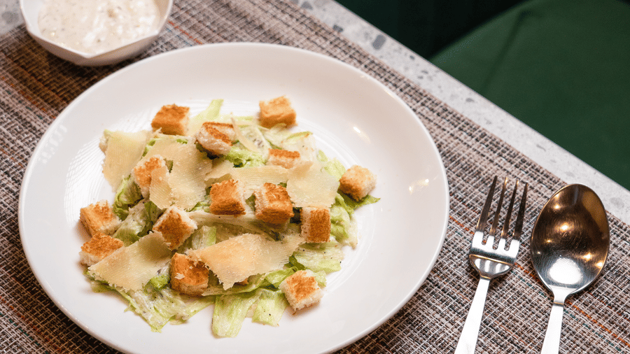 A top-down shot of a fresh Caesar salad with golden croutons and shaved parmesan on a white plate, served with silver cutlery at Apartel by Aarin, Oragadam.