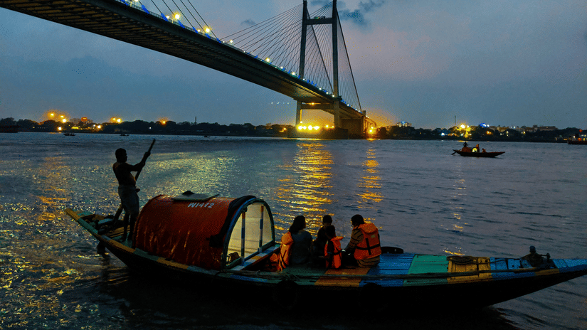 A boat with people on it travelling on a water body in front of the Howrah Bridge in Kolkata during twilight hour.