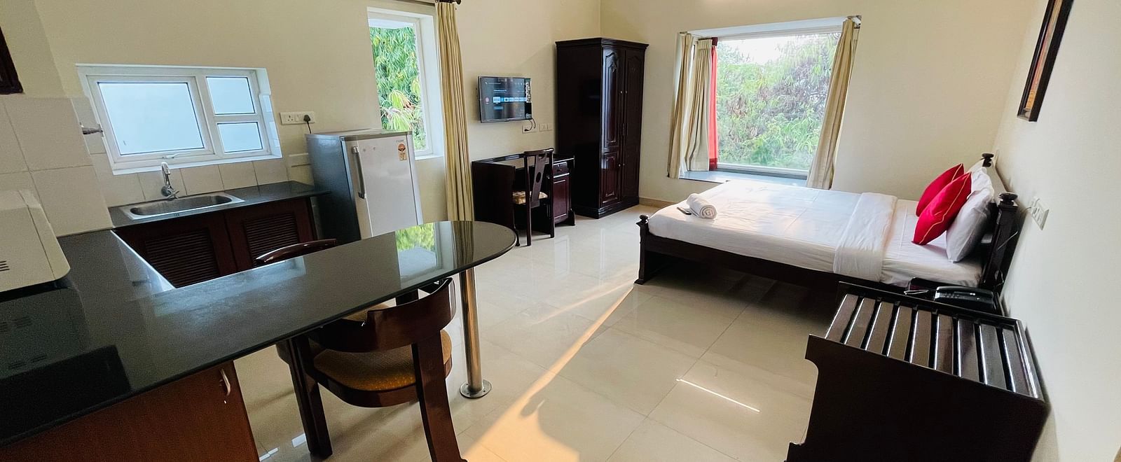 A studio room featuring a double bed with red accents, a desk, a chair, a kitchenette with dining table, chair, and a refrigerator at Sanctum Suites Indiranagar, Bangalore.