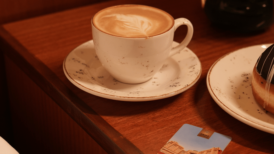 A latte in a white cup sits on a wooden nightstand beside a bed, with a hotel keycard and a vase of purple and white flowers, conveying a cosy morning.
