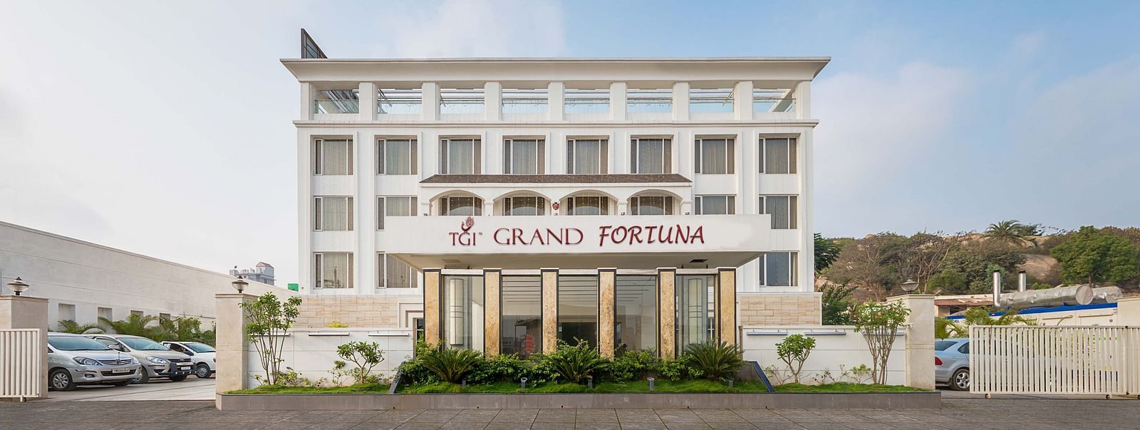 The front exterior of the TGI Grand Fortuna hotel with parked cars and a clear blue sky above - TGI Grand Fortuna, Hosur