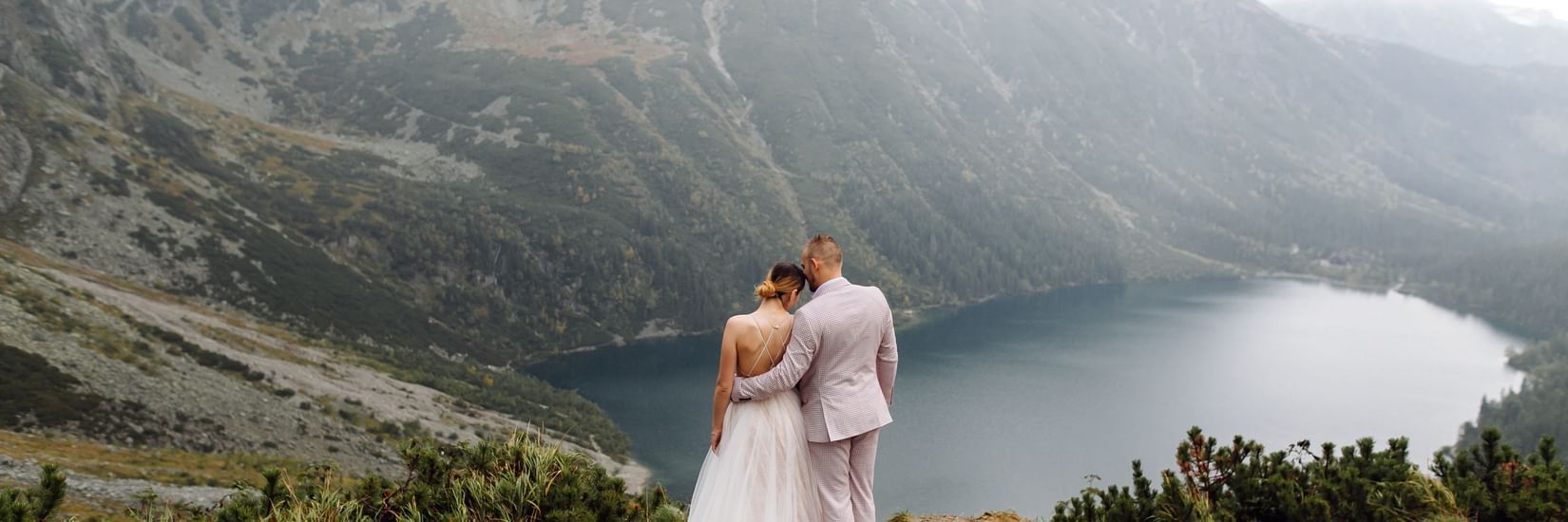 A wedding couple hugging each other on top of a mountain overlooking a lake by the valley.