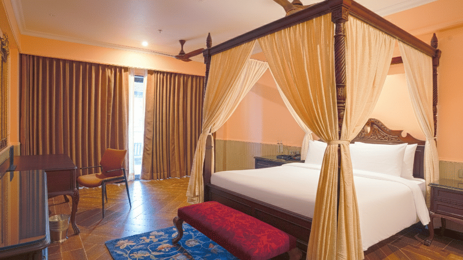 A bedroom with a 4-poster bed draped in sheer curtains, wooden flooring, cushioned bench, sliding curtains and a small dining table set near the window at MAYFAIR Bay Resort, Paradeep.