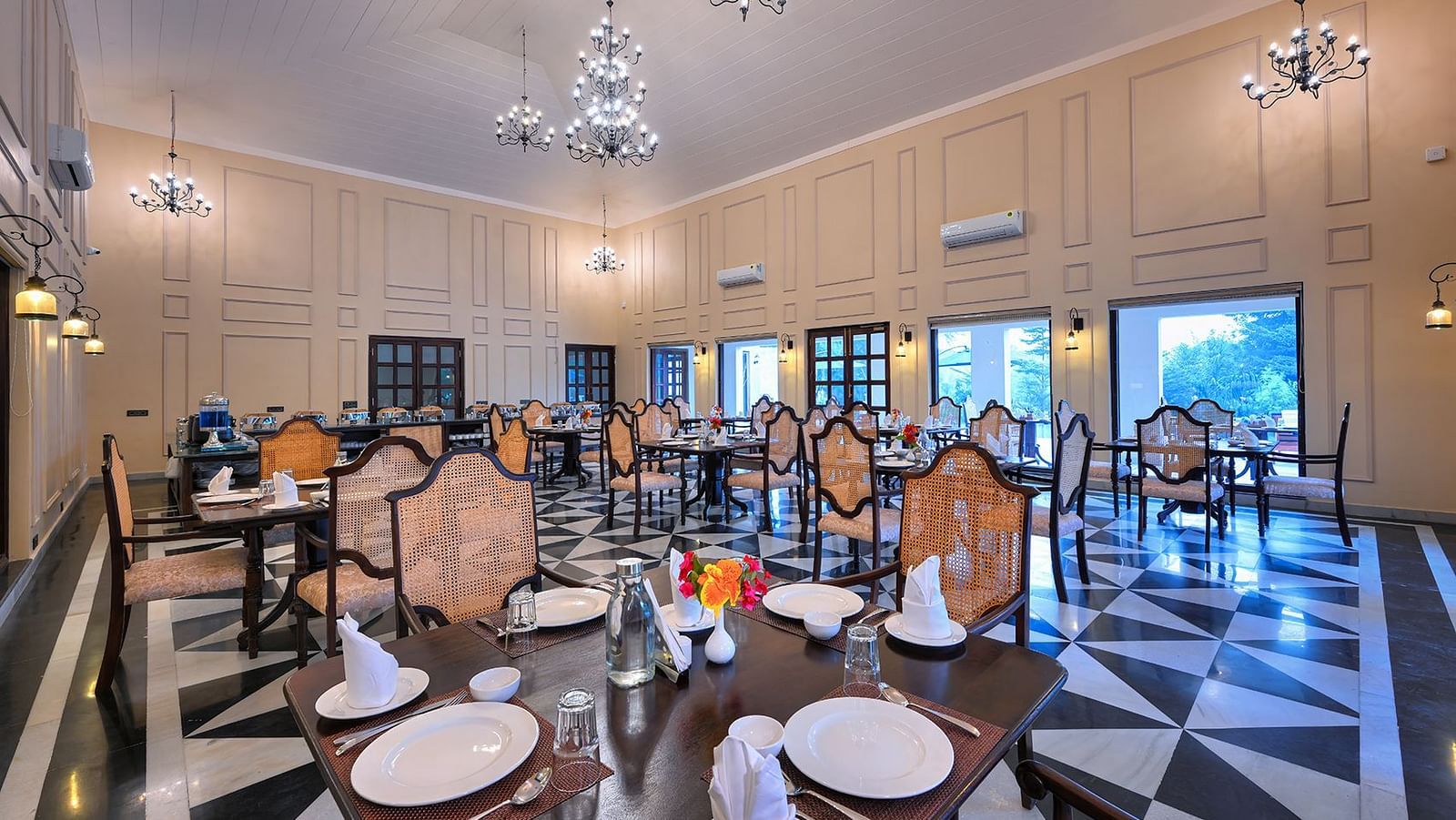 Spacious dining area featuring high ceilings, chandeliers, and classic wooden furniture. The black-and-white patterned flooring contrasts with the warm beige walls, complemented by large windows and elegant wall sconces - Singhvilas, Ranthambore