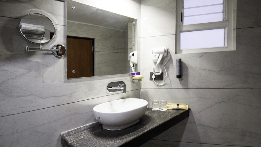 A ceramic washbasin on a dark vanity top, a wall mirror, a hair dryer, and a small window at Valmere Lungta Boutique Hotel & Spa, Darjeeling.