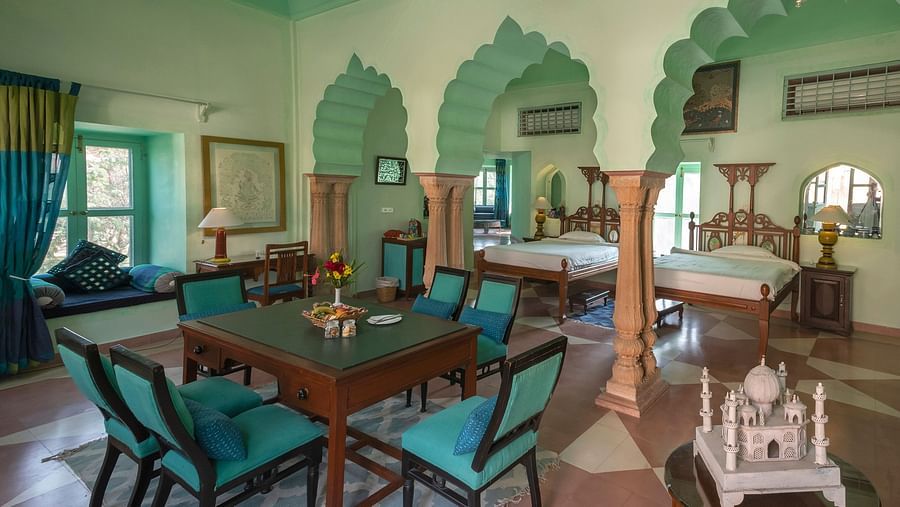 A spacious room at Neemrana's Tijara Fort Palace features a dining table with teal chairs, two beds, arched doorways, and mint green accents.