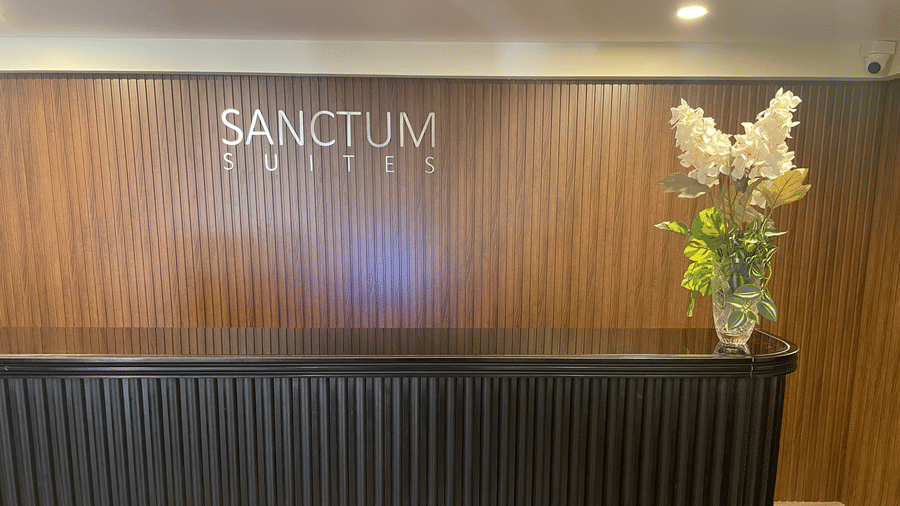 Reception area of Sanctum Suites, Richmond Road, Bangalore features a black counter, a wood panelled wall with signage, and a vase of flowers.