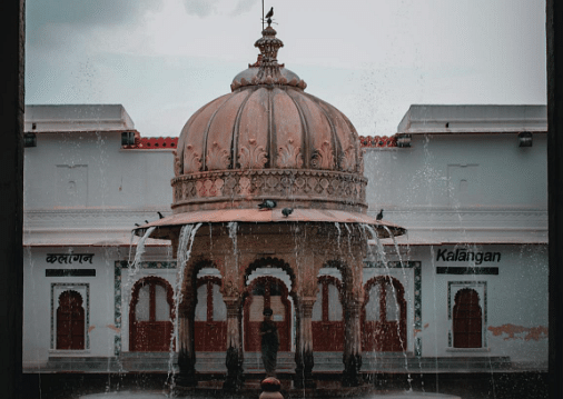 A building with a central dome and flowing water feature, framed by an arched doorway, with text on either side.