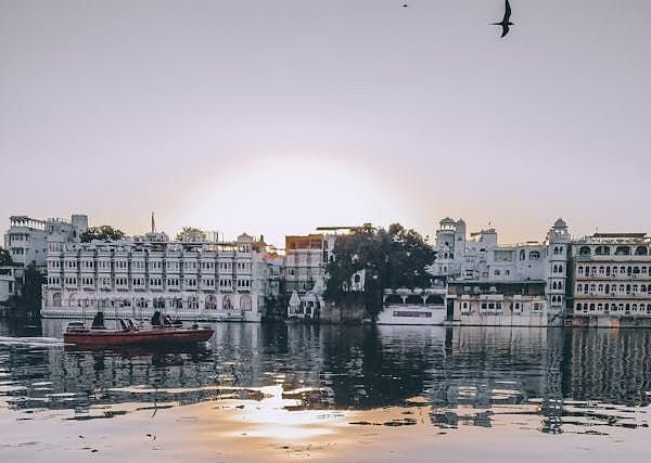 A boat floats on a calm lake reflecting the setting sun, with buildings lining the distant shore.
