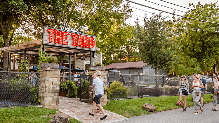 Man walking into a place with a board titled the yard while a group of women walk through a paved road.
