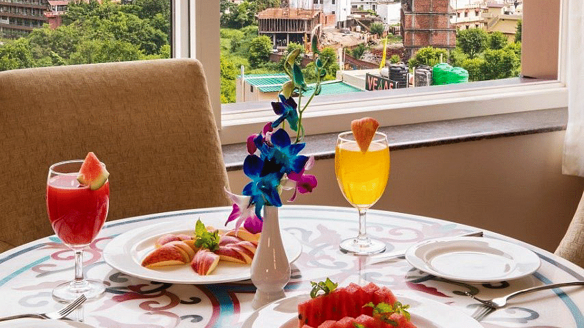 A close up shot of the dining table with well plated fruits and juices overlooking the lush green views from the window at Regenta Inn DLS.
