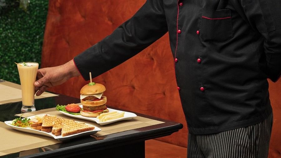 A professional chef wearing a black uniform with red piping and a tall white hat is placing a tall glass of beverage on a table set with a burger and fries. A green plant wall and orange upholstered seating are in the background.  - Clarks Inn Express, Nea