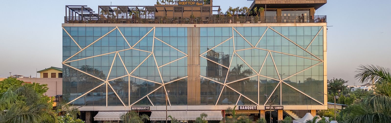 Exterior view of Hotel Highway King, Jaipur, with a large glass facade and rooftop signage, surrounded by greenery.