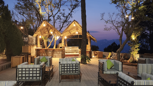 Outdoor seating area at dusk with cushioned chairs and sofas, illuminated by hanging lights under thatched-roof structures, surrounded by trees and a scenic mountain view in the background - Suryavilas Luxury Resort and Spa