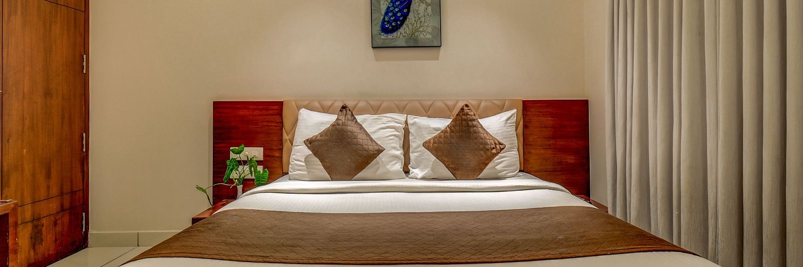 A spacious room at Harigovinda's JNStay with a large bed, wooden headboard, bedside table, curtains pulled to the side, and a photo frame above the headboard