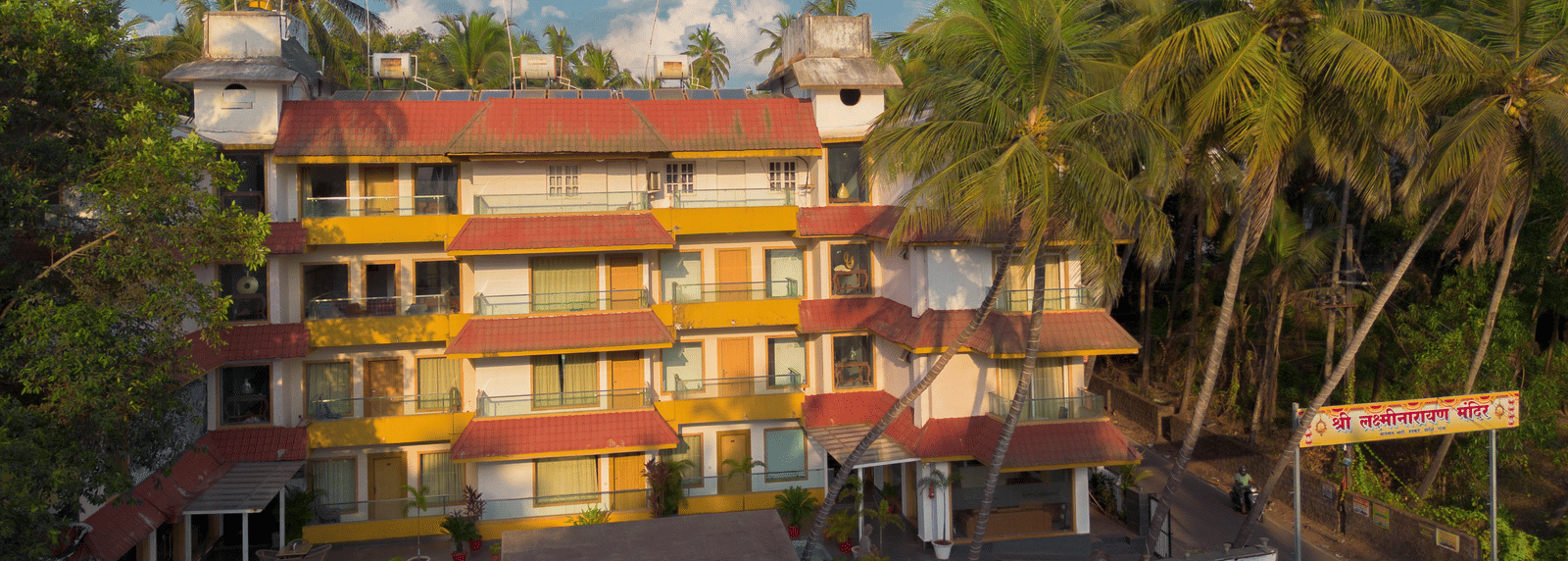 A colourful, low-rise hotel complex with many windows, surrounded by lush green palm trees under a bright sky - Perfectstayz Premium Goa Continental