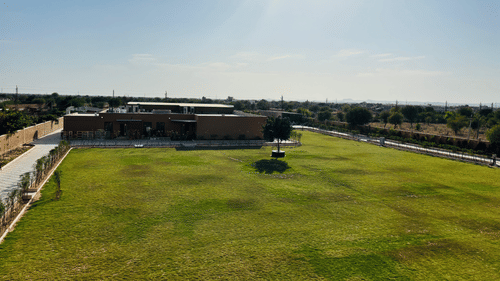 A wide green lawn with scattered trees and structures under bright sunlight at Pride Resort, Jodhpur in the distance.