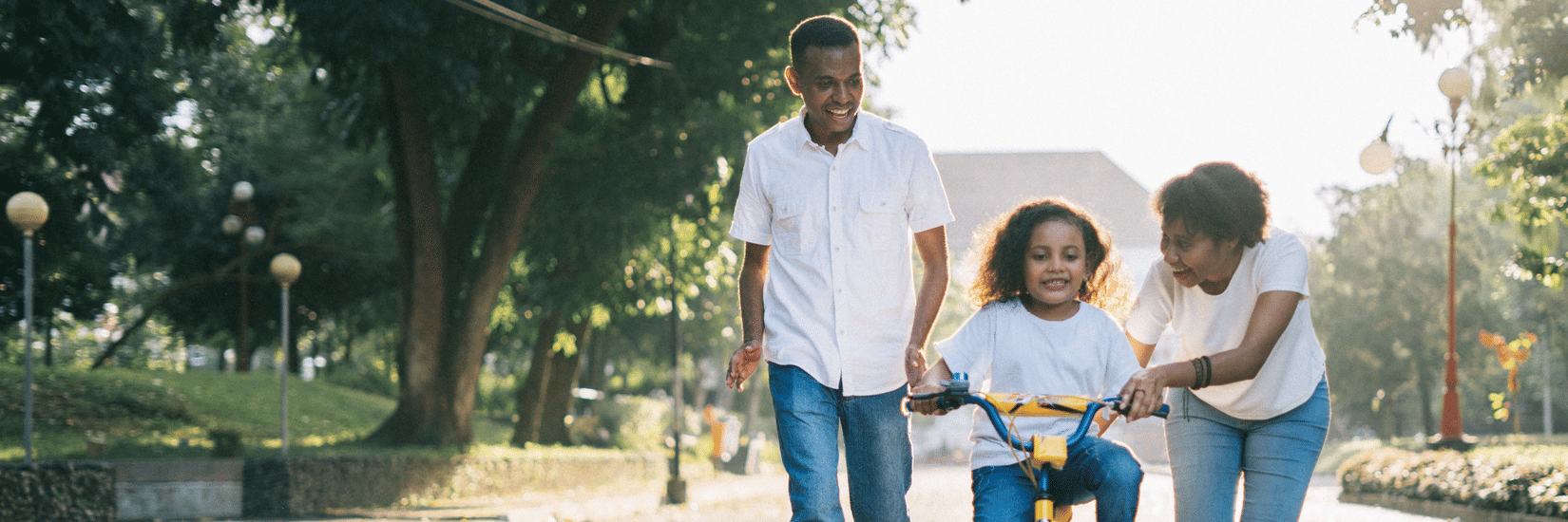 A mother and father smiling as they help their young daughter learn to ride a bicycle with training wheels on a sunny park path.