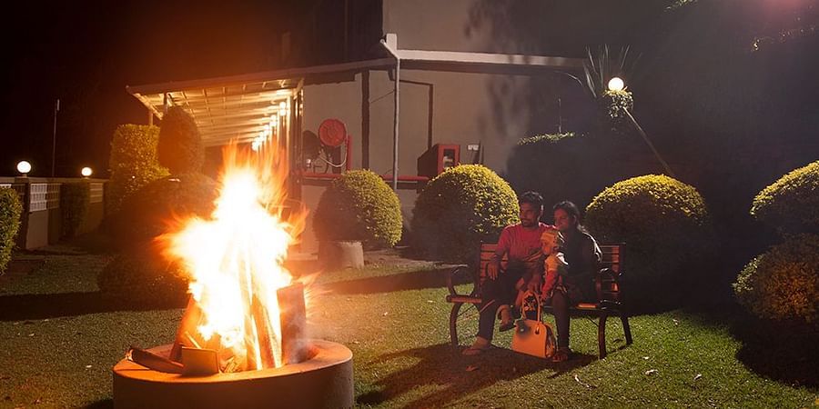 Outdoor bonfire at night. Flames rise from a fire pit, casting light on nearby bushes and a building in the background - Abad Copper Castle, Munnar